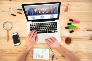 Dłonie piszą na klawiaturze laptopa. Na ekranie napis BLOG. Wokół kolorowe zakreślacze, komórka, herbata.