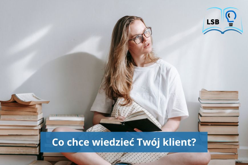 Kobieta o blond włosach, rozpuszczonych, w okularach, zapatrzona na bok, siedzi na podłodze i czyta książkę, obok niej kilka stosów książek.